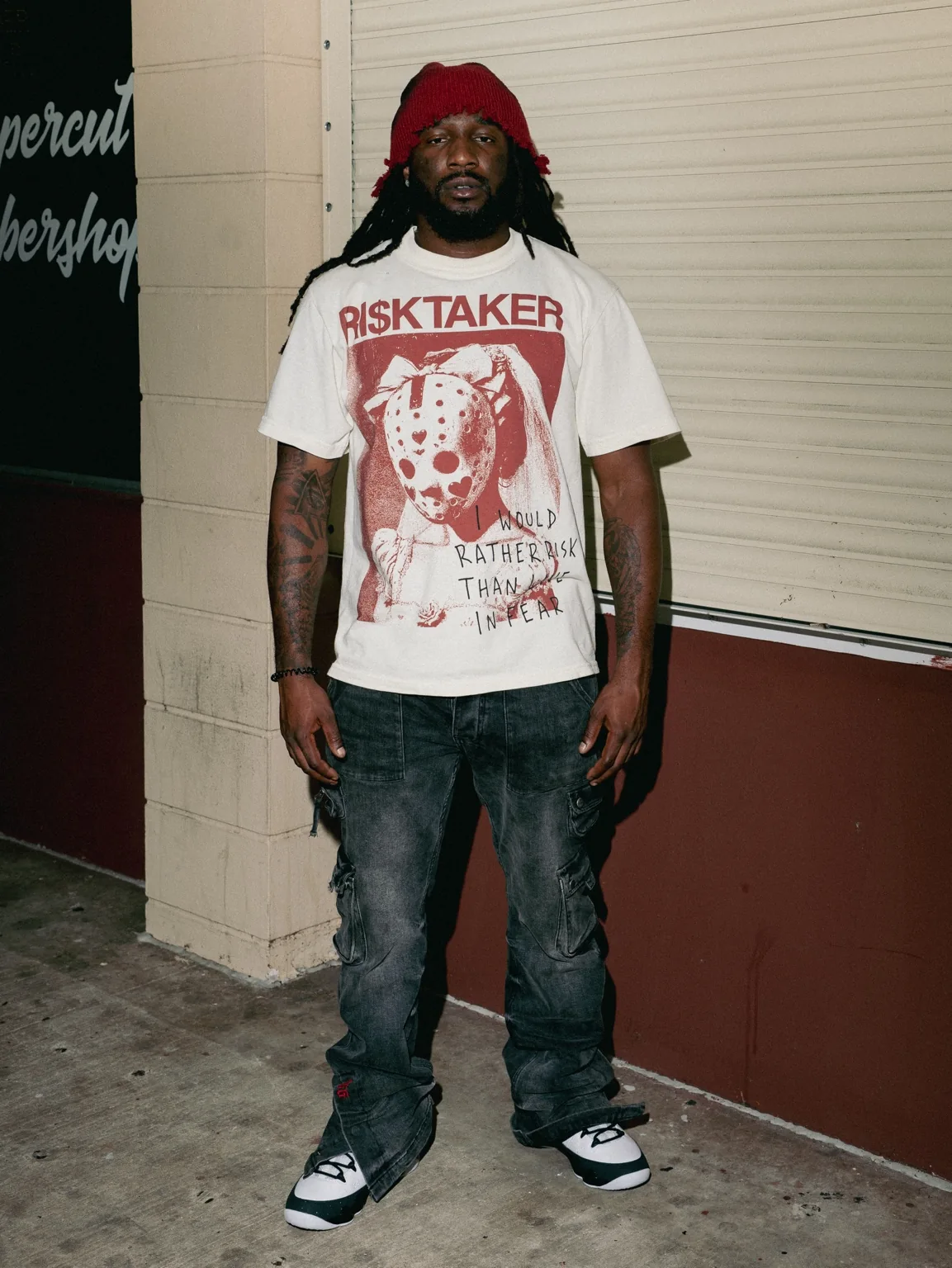 Full-length front view of model wearing the cream RI$KTAKER Mask Tee and charcoal cargo pants